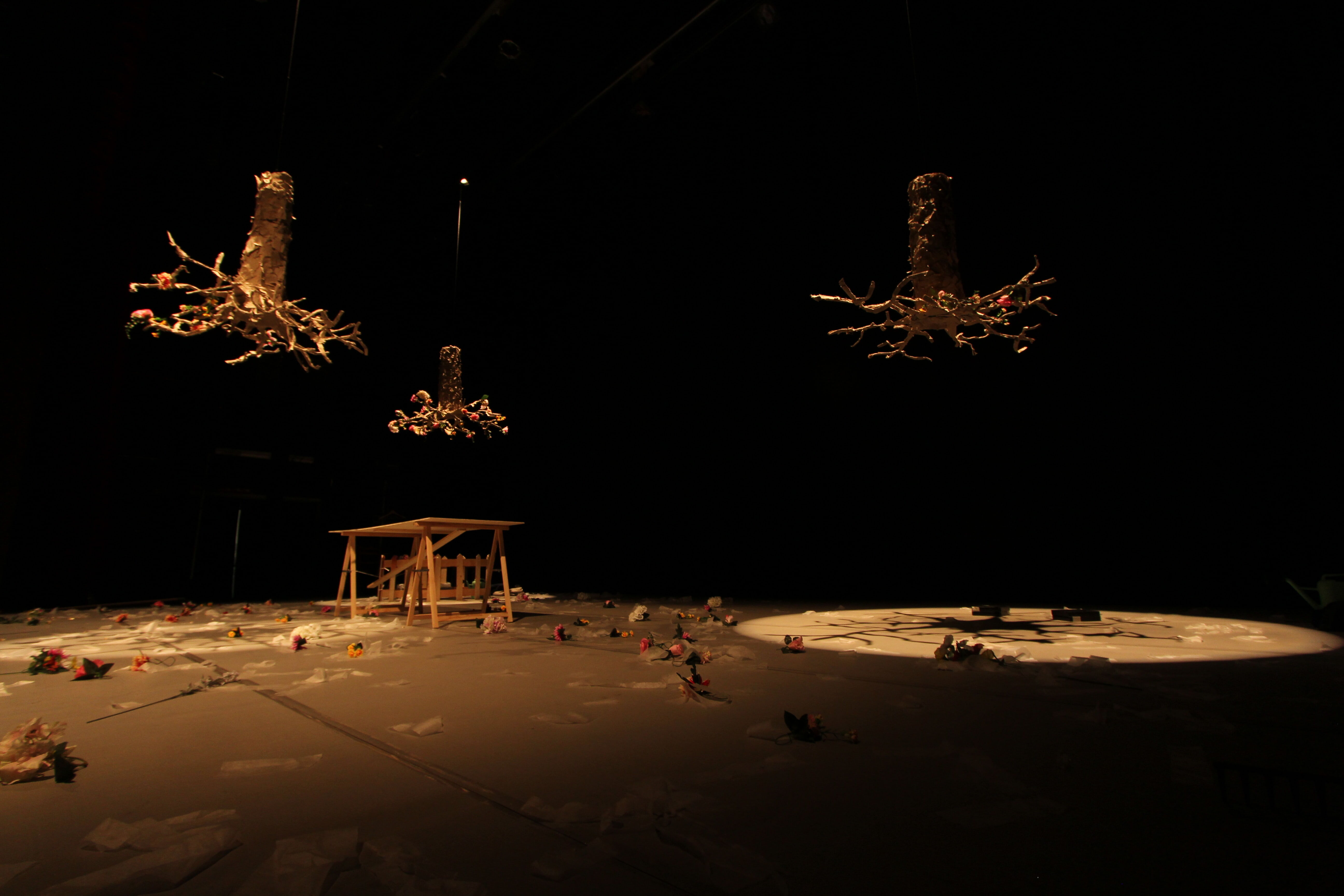 Scenografia di Chicco Margaroli sul palco dello spettacolo Bian o la teoria dei barattoli di Palinodie: tre fusti di alberi finti con rami di varia lunghezza sono appesi a testa in giù, sorretti da cavi trasparenti. Sulla sinistra un tavolo da lavoro spoglio, a terra sono sparsi vari fiori finti colorati e carta velina.