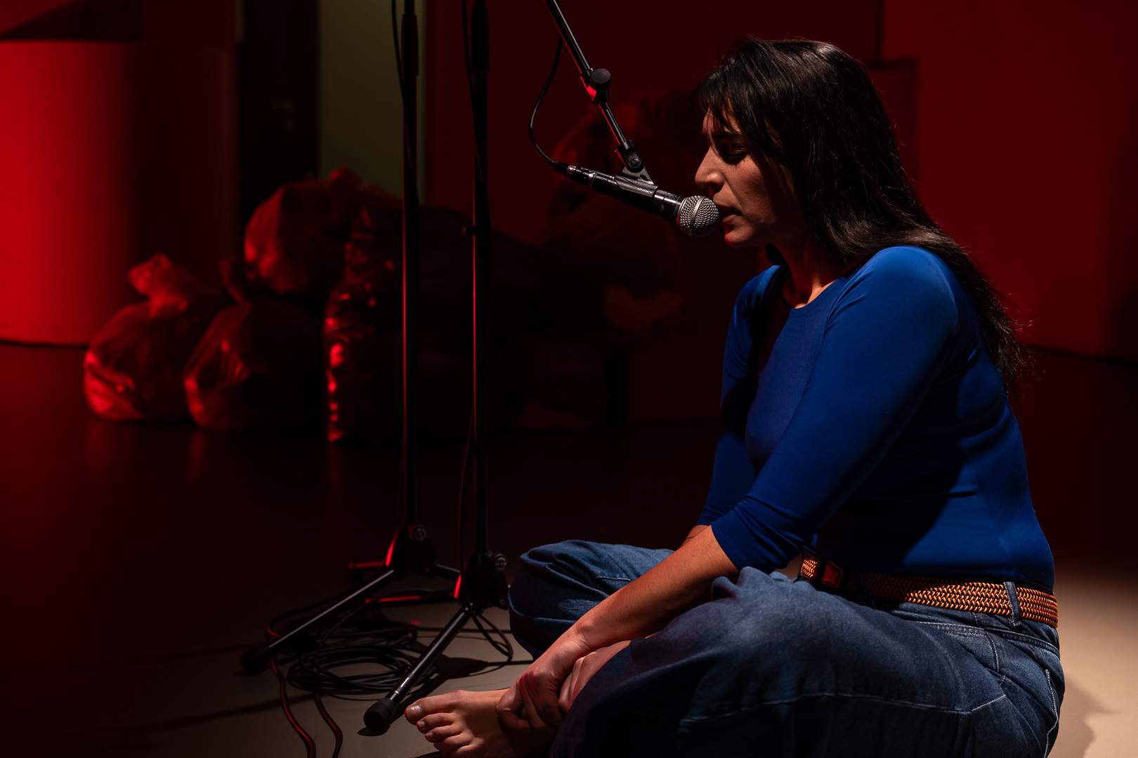 Attrice Silvia Pietta seduta in primo piano recita al microfono su sfondo rosso nello spettacolo sulla violenza donne Come noi