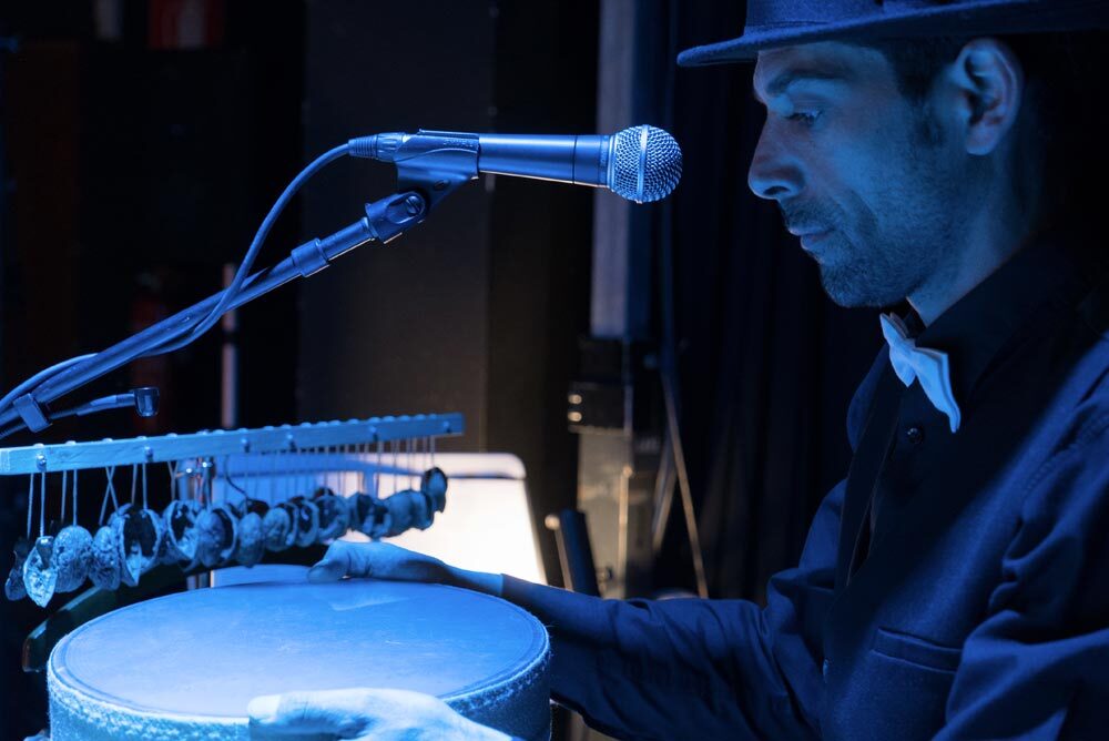 Luca Gambertoglio rumorista live in Un Raconto di draghi si vede di profilo destro mentre è intento a tenere tra le mani un tamburo e in posizione frontale al microfono di scena. In secondo piano una fila di conchiglie sospese ad un travetto.
