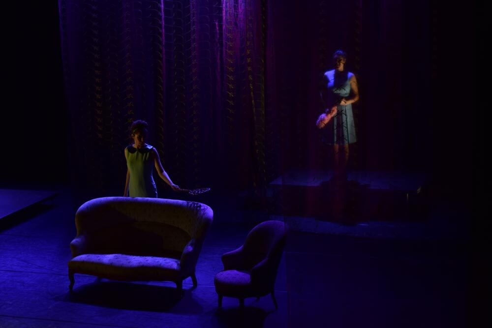 Mariachiara Caneparo attrice in primo piano sul palco si intravede illuminata di blu, Giulia Sartore attrice alle sue spalle è celata da un tenda velata di scena in Donne che si vogliono sposare. Forse